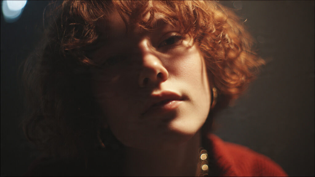 A close-up, moody portrait of a woman with curly red hair and a thoughtful expression. Golden light filters across her face, casting soft shadows against a dark background.