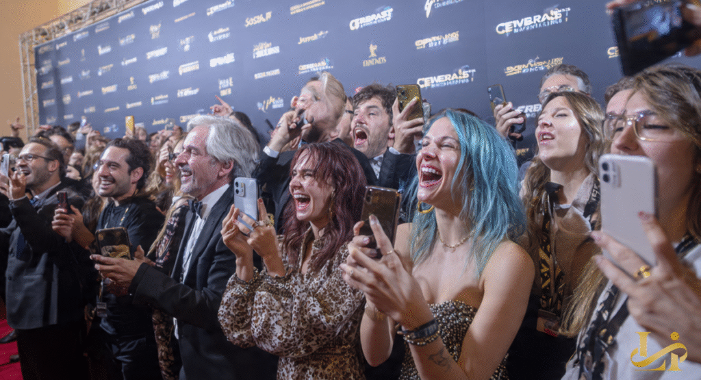 A crowd of enthusiastic people on a red carpet, many holding up smartphones to capture an event, laughing and cheering. The scene captures intense fan reactions and celebrity responses during a glamorous occasion.