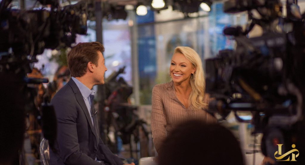 A wide shot of a morning talk show set featuring a guest and a host engaged in a cheerful, laughing conversation. The guest has long blonde hair and wears a striped blouse, while the host sits in profile on the left.
