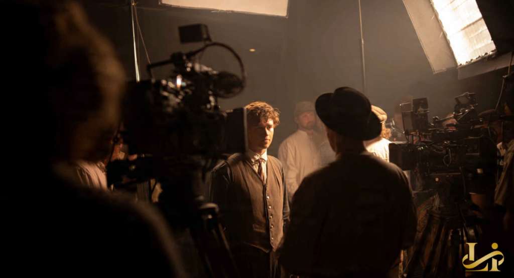 An actor in period costume stands on a film set, illuminated by bright production lights and surrounded by crew and equipment. This depicts a pivotal moment in a breakthrough role, likely as Ledger Ward.