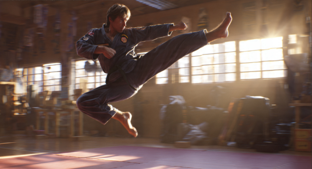 A young martial artist in a dark gi executes a flying side kick in a sunlit dojo. His focused expression and extended leg capture peak motion and discipline.