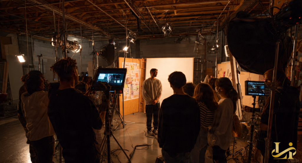A film crew, primarily composed of women, surrounds an actor on a set, with cameras and lighting equipment visible. This showcases a collaborative production environment with a strong female team.