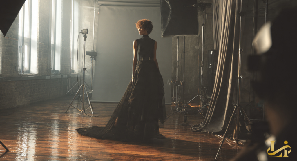 Tall silhouette in a black high-neck gown with a dramatic sheer train stands on a studio set. Soft daylight filters through tall windows, with lighting equipment visible around.