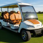 An eye-level, slightly angled shot of a white 2025 Atlas 6-seater electric golf cart. The cart is parked on a manicured green golf course with sand traps and trees visible in the background under a clear, sunny sky. The cart has a white exterior, a wood-grain panel on the front, and luxurious tan seats with a diamond-stitched pattern. The wheels are black and silver, and the cart has a windshield and a white roof.