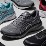 A flat lay product photograph of several pairs of ASICS running and training shoes arranged on a light gray surface. The shoes are in various colors, including light blue, gray, red, and black, highlighting a range of models.