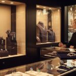 Two women in a luxury jewelry store. A woman on the right, presumably a sales associate, holds a tray with a diamond and sapphire brooch, presenting it to a customer on the left. Display cases filled with elegant necklaces, earrings, and rings with sapphires and diamonds are visible in the foreground and a larger, lit-up case in the background. The store's interior is sophisticated, with a view of a sunny Beverly Hills street outside.