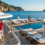 An aerial view of the deck of a luxury yacht with an infinity pool overlooking a clear blue sea and towering cliffs. Guests in white and red outfits are socializing on lounge chairs and around a dining table, illustrating a luxury cruising experience.