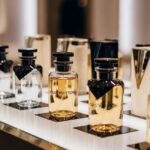 A close-up display of multiple Louis Vuitton fragrance bottles, alternating between clear glass with amber liquid and dark bottles, set on a bright white and black surface in a retail setting.