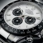 A detailed, angled close-up photograph of a Rolex Cosmograph Daytona "Panda" watch. The timepiece features a white dial with three contrasting black sub-dials, a black Cerachrom bezel with a tachymeter scale, and a polished stainless steel Oyster bracelet.