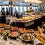 A bright, spacious dining hall on a cruise ship or a restaurant with a panoramic view of the ocean through large windows. A long, central buffet table is laden with an abundant and artistically arranged variety of dishes, including salads, cold platters, breads, and various seafood and meat dishes. Guests are seated at well-appointed tables with white tablecloths, and waitstaff in black vests are attending to them, creating an atmosphere of elegant dining.