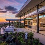 A wide exterior view of a modern luxury home at sunset. The home features a large, open-air patio with white lounge furniture and an outdoor garden area. The interior is visible through floor-to-ceiling glass doors, showcasing a contemporary kitchen and bedroom. The home overlooks a scenic landscape with hills and the ocean in the distance.