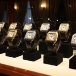 A professional photograph of a collection of Cartier Santos watches displayed in a glass case. The watches, featuring different case sizes, materials, and strap options, are arranged on black stands, highlighting their iconic square shape and visible screws.