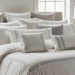 A luxuriously made bed filled with a variety of white and gray pillows. The pillows are arranged in a layered, inviting fashion, some with subtle patterns and textures. The bedding is also white with a gray, textured blanket draped over the foot of the bed.