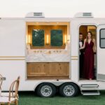 A professional photo of a modern, multi-stall luxury restroom trailers parked at a sophisticated outdoor event venue. The sleek exterior of the trailer is a dark color, and a partial view inside reveals high-end features like vanity mirrors, good lighting, and clean countertops, emphasizing comfort and style.