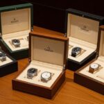 An open, elegant wooden watch box showcasing a collection of ten different luxury watches. The watches are arranged neatly on soft, individual cushions, with the box's plush interior lining and polished brass hinges visible. The watches feature various styles, including chronographs, divers, and classic dress watches, with different straps and metal finishes. The overall image conveys a sense of luxury, organization, and careful curation.