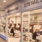 A view of a modern and clean nail salon with large front windows. The interior shows several clients getting manicures and pedicures in luxurious brown leather chairs. Signs for "The Nail Bar" and "Elite Nail Studio" are visible on the exterior.