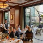 An elegant yacht club restaurant with a retractable glass wall opening to a city terrace. Guests are seated at fine dining tables, being served by waiters, while a jazz band performs on a small stage in the corner.