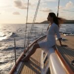 A young woman, presumably Ashley Marti from Below Deck Sailing Yacht Season 3, sits barefoot on the teak deck rail of a sailboat, wearing a white dress, with her hair blowing in the wind as she gazes out at the ocean during a sunset.