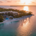An aerial view of Baker's Cay Resort Key Largo at sunset, showcasing the modern main building and expansive beachfront lined with palm trees and sun loungers. The golden light reflects off the calm turquoise water of the ocean, highlighting the exclusive beach escape.