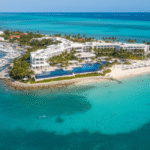 An aerial view of the luxurious Blue Haven Resort in Turks and Caicos, situated on a narrow peninsula with a white-sand beach, infinity pool, and an adjacent marina filled with yachts. The resort is surrounded by the brilliant turquoise water of the Caribbean Sea.