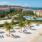An aerial view of Bolongo Bay Beach Resort on St. Thomas, showing the colorful hotel buildings, a large swimming pool, a beach volleyball court, and a white-sand beach dotted with hammocks, lounge chairs, and palm trees, with guests relaxing and swimming in the turquoise water.