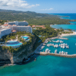 An aerial view of the El Conquistador Resort in Puerto Rico, dramatically situated on a cliff overlooking a large marina filled with boats. The resort features white buildings, pools, and is surrounded by lush green hills and the turquoise Caribbean Sea.
