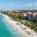 An aerial view of the Casa Marina Key West, Curio Collection resort, showing its stately white buildings with red tile roofs along a long, wide stretch of pristine white-sand beach. The turquoise ocean meets the shore, with palm trees and a pool area on the resort grounds, and the Key West skyline visible in the distance.
