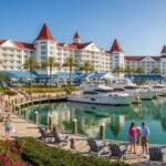 An exterior view of Disney's Yacht Club Resort, featuring a large white and red-roofed building overlooking a marina with several yachts docked, surrounded by lush green lawns, pink flowers, and palm trees, with guests walking along the boardwalk.