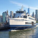 A large white and blue ferry boat labeled "GOVERNORS ISLAND" docking at a pier with a crowd of people waiting to board or disembark. The New York City skyline, including the Freedom Tower (One World Trade Center), is clearly visible in the background under a sunny blue sky.