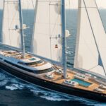 An aerial view of the massive three-masted sailing superyacht Koru, reportedly belonging to Jeff Bezos and valued at $500 million, sailing on the ocean with all white sails deployed, and a helicopter flying alongside it, showcasing its deck pool and vast size.