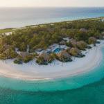 An aerial view of Little Palm Resort, a secluded, private tropical island in the Florida Keys, featuring thatched-roof bungalows, a central pool, lush palm trees, a white-sand beach, a small airstrip, and yachts docked at the piers, all surrounded by clear turquoise water.