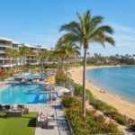 An inviting view of the San Diego Mission Bay Resort, showing a modern white hotel building, a large swimming pool lined with palm trees, and a sandy beach leading to the calm waters of Mission Bay, with a paddleboarder visible in the distance.