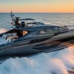 An aerial side view of a sleek, dark grey and black Tecnomar for Lamborghini 63 motor yacht speeding across the ocean at sunset, creating white wake on the dark blue water.