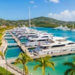 An aerial view of the Yacht Haven Grande Marina in St. Thomas, showcasing a bright, luxurious coastal oasis with rows of large, white superyachts docked in the turquoise water, palm-tree-lined promenades, and luxury waterfront buildings against a backdrop of green, rolling hills.