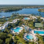 An aerial view of the Sandpiper Bay resort complex in Port St. Lucie, Florida, showing multiple large swimming pools, extensive tennis courts, hotel buildings, and a marina along a wide, deep blue river or intercoastal waterway surrounded by lush suburban greenery.