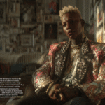 A high-fashion portrait of Miss J Alexander wearing an ornate, sparkling gold and pink brocade blazer. He is seated in a room filled with framed photos, looking directly at the camera with a poised expression.