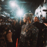 A woman with sleek hair and red lipstick wearing a vibrant, multi-colored metallic gown. She looks directly into the camera lens while soft red and orange lights glow in the background.