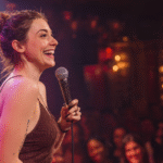A woman with a messy bun laughs joyfully while holding a microphone on a comedy club stage. She wears a brown textured top as the warm stage lights glow behind her.