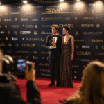 A man in a tuxedo holding an award and a woman in a sparkling gown pose on a red carpet, surrounded by photographers and a backdrop with multiple "CÉSAR" logos. The scene captures a public appearance, where a celebrity thanks his sublime companion at France's César Awards.
