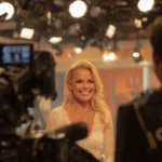 A professional television studio setting showing a blonde woman smiling warmly during a live broadcast. Bright studio lights and cameras are visible in the foreground, framing her as the central subject of the interview.