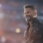 A close-up portrait of a smiling man with a groomed beard and styled hair wearing a sequined black jacket. He is standing in a brightly lit stadium environment.