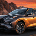 A dark bronze 2026 Toyota Highlander parked on a scenic mountain road during a vibrant orange sunset. The vehicle features sleek LED headlights and a prominent front grille reflecting the golden hour light.