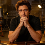 A man in a black polo rests his chin on clasped hands at a wooden table with film reels, books, and painted portraits on easels behind him.