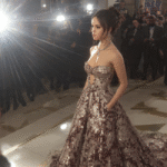 Jenna Ortega walks the red carpet in a stunning, shimmering, strapless gown with a full skirt. She is surrounded by photographers and bright camera flashes, capturing her elegant red carpet moment.