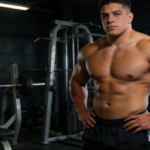 Joseph Baena stands shirtless in a gym with hands on hips. A vintage bodybuilding poster hangs behind him near the rack.