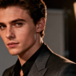 A striking headshot of Nicholas Galitzine, looking intently at the camera in a dark suit. A studio spotlight glows in the blurred background, highlighting his captivating presence.