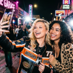 Two young women smile broadly while taking a selfie on a crowded city street at night. Alabama Barker wears a colorful racing-style jacket while surrounded by paparazzi and glowing neon signs.