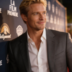Alexander Skarsgård smiles beside a step-and-repeat at an L.A. film premiere. Photographers’ flashes glow behind him as he wears a grey suit and open white shirt.