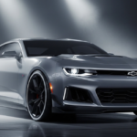 A sleek, silver performance coupe sits under bright studio spotlights, showcasing a futuristic front fascia and aerodynamic body lines. This conceptual design represents the highly anticipated Chevy Camaro Replacement featuring advanced LED lighting and a wide, aggressive stance.