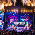 A wide-angle view of a massive stadium stage with pyrotechnics exploding as two performers stand under a giant Chris Brown and Usher Tour logo.